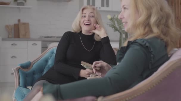 Δύο χαρούμενες ώριμες λευκές γυναίκες που κάθονται σε αναπαυτικές πολυθρόνες κρατώντας χρήματα και πούρα και μιλώντας. Παλιές φίλες κουτσομπολεύουν για τη ζωή τους. — Αρχείο Βίντεο