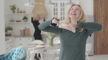 Elinde puro, etrafa para saçan ve gülümseyen zengin, olgun, beyaz bir kadın. Olumlu arkadaşı arka planda şarap ve bardaklarla geziyor. Lüks mutfakta dinlenen iki zengin yaşlı bayan..