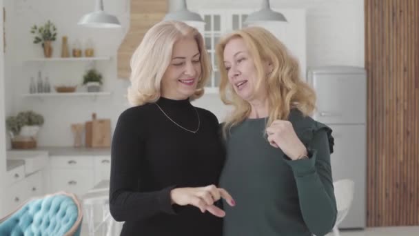 Посміхаючись, зрілі кавказькі жінки стоять у розкішній кухні і стискають пальці у формі серця. Багаті старші жінки проводять разом вільний час.. — стокове відео
