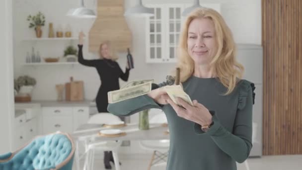 Reiche reife kaukasische Frau, die Geld verstreut und lächelt. Ihr positiver Freund tanzt mit Wein und Gläsern im Hintergrund. zwei wohlhabende Seniorinnen ruhen sich zu Hause aus. — Stockvideo