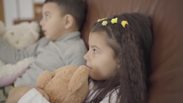 Cara de cerca de una linda chica de Oriente Medio sentada con su hermano en el entrenador y sosteniendo un oso de peluche. Pequeña dama sonriente pasar la noche en casa acogedora con la familia . — Vídeos de Stock