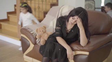 Orta Doğulu bir anne koltukta oturuyor ve iki küçük çocuğu etrafta koşuşturuyor. Güzel bir kadın elleriyle başını tutuyor ve tatlı kızı yukarı geliyor..