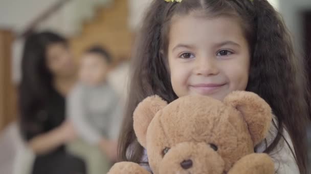 Close-up face of a happy Middle Eastern girl with brown eyes and curly hair holding the teddy bear and smiling. Child spending evening at home with her brother and mom sitting at the background. — Stock Video