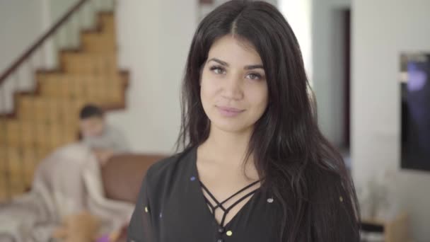 Beautiful Middle Eastern woman with hazel eyes and long dark hair looking at the camera and smiling. Her two little children playing at the background in cozy home. Happy family. — Stock Video