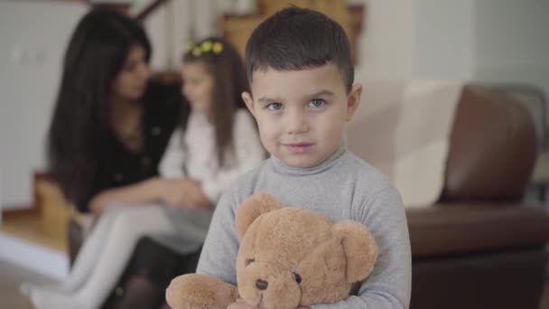 Charmanter Junge aus dem Nahen Osten mit grauen Augen und dunklen Haaren, der den Teddybär hält und in die Kamera schaut. Kind posiert zu Hause, während seine Schwester und Mutter im Hintergrund spielen. — Stockvideo