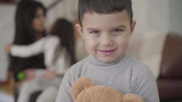 Cara de cerca de un encantador chico de ojos grises de Oriente Medio con el pelo oscuro sosteniendo al oso de peluche y mirando a la cámara. Niño posando en casa mientras su hermana y mamá juegan en el fondo . — Vídeo de stock