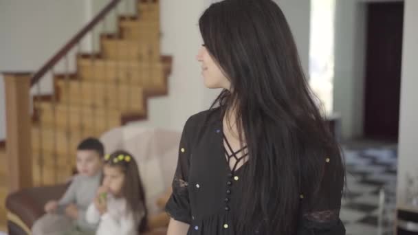 Sorridente donna del Medio Oriente con gli occhi nocciola e lunghi capelli scuri guardando i suoi figli seduti sullo sfondo e tornando alla fotocamera. Felice madre passare del tempo con suo figlio e sua figlia . — Video Stock