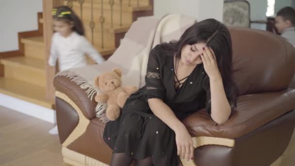 Tired Middle Eastern mother sitting on the armchair and her two little children running around. Beautiful woman holding the head with hand and her cute daughter coming up. — Stock Video