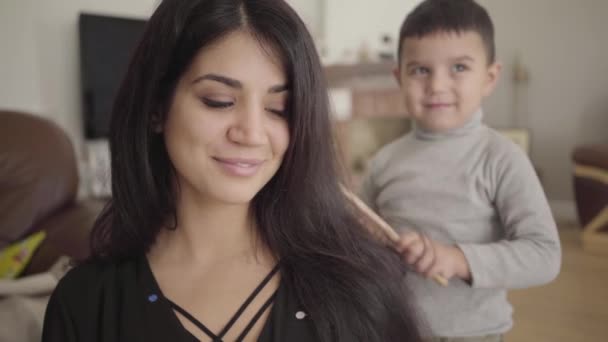 Retrato de uma mulher de olhos castanhos do Oriente Médio penteada pelo menino sorridente que fica no fundo. Filho se preocupando com sua adorável bela mãe . — Vídeo de Stock