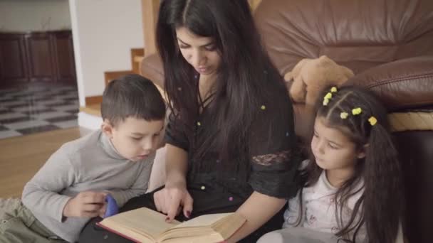 Γοητευτική γυναίκα από τη Μέση Ανατολή που διαβάζει το βιβλίο σε μικρά παιδιά. Μητέρα που εκπαιδεύει τον αγαπημένο της γιο και κόρη στο σπίτι. — Αρχείο Βίντεο