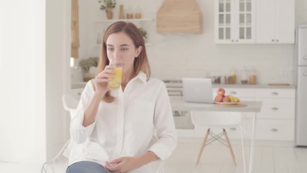 Joven hermosa mujer caucásica sentado en la luz moderna cocina bebiendo jugo de naranja e ir al fondo de la computadora portátil. Chica bastante sonriente pasar los fines de semana en casa . — Vídeo de stock