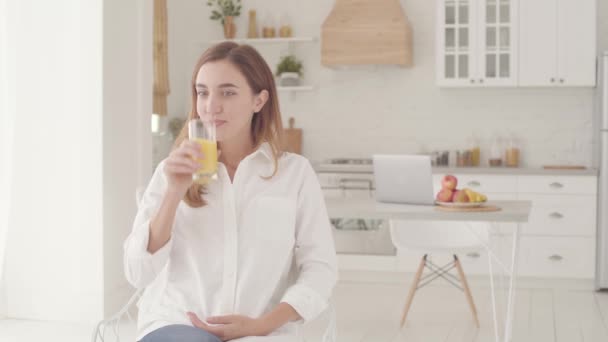 Portret van een jonge mooie blanke vrouw zittend in een lichte moderne keuken en sinaasappelsap drinkend. Vrij lachend meisje brengt het weekend thuis door. — Stockvideo