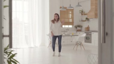 Neşeli, beyaz bir kızın gündelik kıyafetleriyle modern mutfakta enerjik dansı. Mutlu güzel kadın hafta sonları evde eğleniyor..
