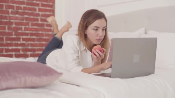 Młoda biała kobieta leżąca w łóżku, pisząca na laptopie i jedząca czerwone jabłko. Piękna uczennica uczy się w domu w swobodnej atmosferze. — Wideo stockowe