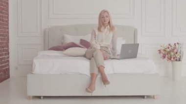 Yatakta dizüstü bilgisayarla oturan ve düşünen gri gözlü sarışın beyaz iş kadınının portresi. Resmi kıyafetli genç bir bayan evden internet üzerinden çalışıyor..