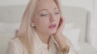 Sarışın, gri gözlü, yorgun ve iç çeken beyaz bir kadının yakın çekim yüzü. Yorgun kadın evde tek başına vakit geçiriyor..