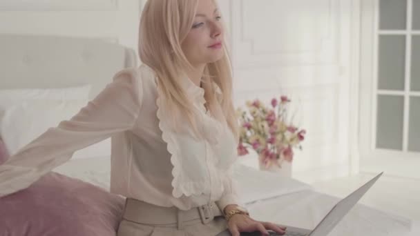 Smiling blond Caucasian businesswoman typing on the laptop sitting on the bed at home. Young beautiful lady in official clothes working online from home. — ストック動画