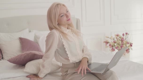 Leende blond gråögd vit affärskvinna som sitter med laptop på sängen och tänker. Ung vacker dam i officiella kläder arbetar online hemifrån. — Stockvideo