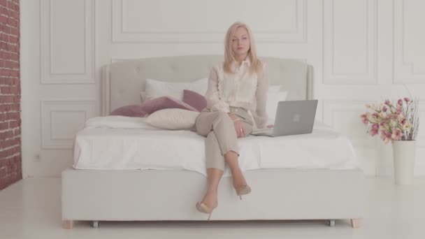 Egy szőke, fehér üzletasszony portréja, szürke szemekkel, laptoppal az ágyon ülve és gondolkodva. Fiatal gyönyörű hölgy hivatalos ruhákban dolgozik online otthonról. — Stock videók