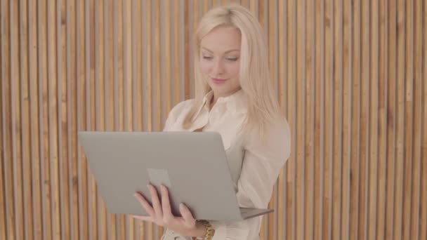 Schöne kaukasische Frau mit blonden Haaren und grauen Augen, die den Laptop hält und eine Siegesgeste macht. selbstbewusste junge Frau auf dem Hintergrund einer Holzwand. — Stockvideo