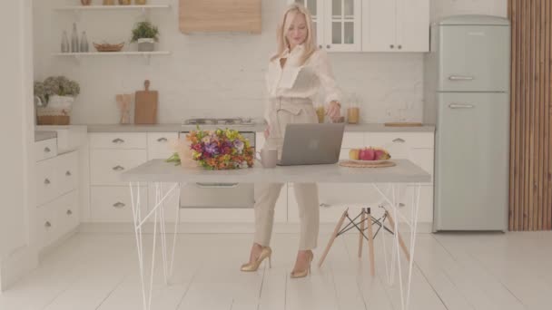 Feliz señora rubia caucásica de pie en la cocina moderna con una taza de té o café y un ramo de flores. Mujer adulta feliz en ropa oficial comenzando un nuevo día con una sonrisa . — Vídeos de Stock