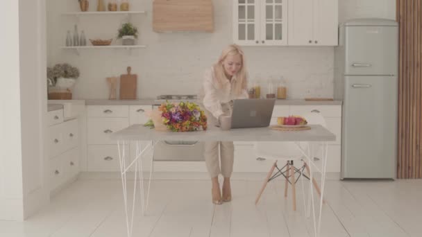 Feliz dama caucásica con el pelo rubio de pie en la cocina moderna, mirando a la computadora portátil y haciendo gesto de victoria. Mujer adulta en ropa oficial empezando a recibir buenas noticias en línea . — Vídeo de stock