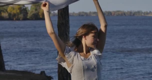 Retrato de una hermosa mujer caucásica en vestido ligero de pie en la orilla con los ojos cerrados sosteniendo bufanda en las manos levantadas. Muchacha atractiva disfrutando del viento soplo desde el mar . — Vídeos de Stock