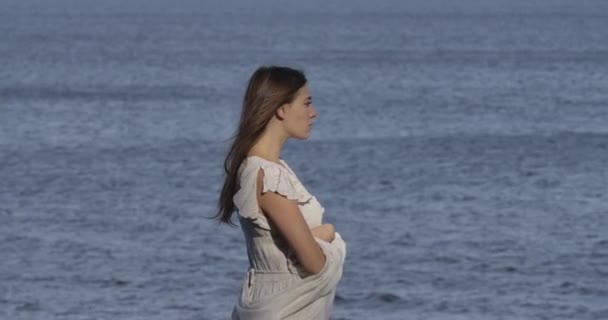 Vue latérale d'une belle femme caucasienne en robe blanche pâle debout sur le fond des vagues de la mer et de la pensée. Attrayant fille passer des vacances sur la plage . — Video