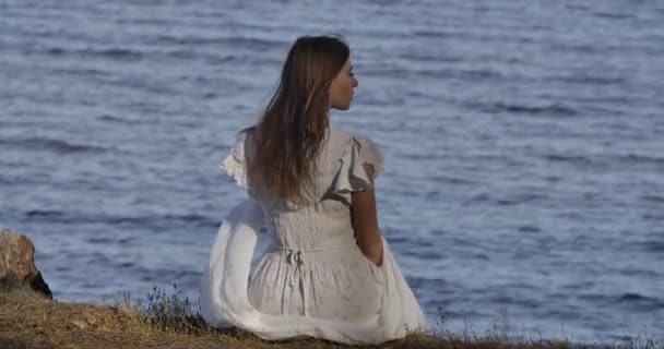 Widok z tyłu eleganckiej białej kobiety w jasnobiałej sukience siedzącej na plaży i myślącej. Piękna dziewczyna spoczywa na tle fal morskich. — Wideo stockowe