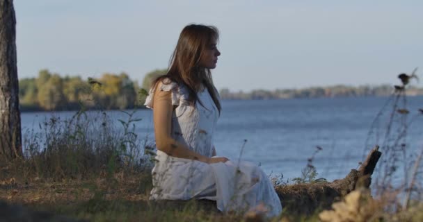 Misteriosa mujer caucásica sentada en la orilla del lago y mirando a su alrededor. Hermosa chica en vestido blanco claro descansando al aire libre . — Vídeos de Stock