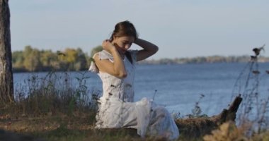 Çekici beyaz kadın yaz gününde gölün kıyısında oturuyor ve saçlarına dokunuyor. Açık beyaz elbiseli güzel kız dışarıda dinleniyor..