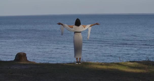 一个漂亮的白种人女人穿着浅白的衣服站在波浪的背景上，双手平整，背向后看。 快乐的女孩在海滩度过夏天. — 图库视频影像