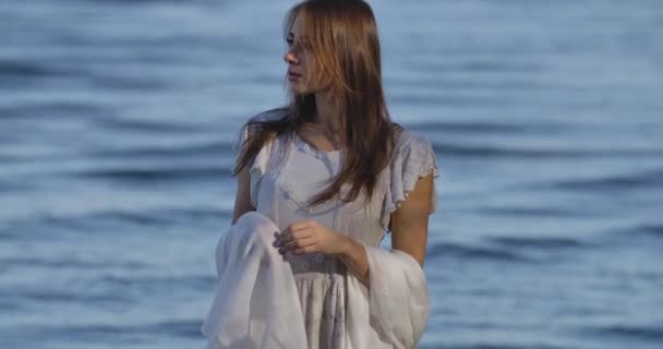 Porträtt av en omtänksam vit kvinna i ljus vit klänning står på bakgrunden av havsvågor. Attraktiv tjej tillbringar sommarkväll på stranden. — Stockvideo