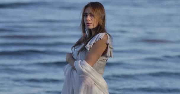 Retrato de una misteriosa chica caucásica en vestido blanco claro de pie sobre el fondo de las olas del mar. Mujer atractiva mirando a la cámara y a un lado — Vídeos de Stock