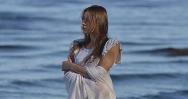 Mystisk vit flicka i ljus vit klänning står på bakgrunden av havsvågor. Vacker kvinna tittar till sidan eftertänksamt. — Stockvideo