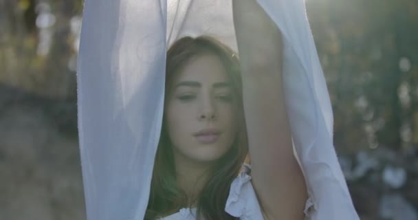 Крупным планом изображена улыбающаяся кавказская девушка, стоящая на солнце с белым шарфом в поднятых руках. Красивая женщина проводит летний вечер на открытом воздухе . — стоковое видео