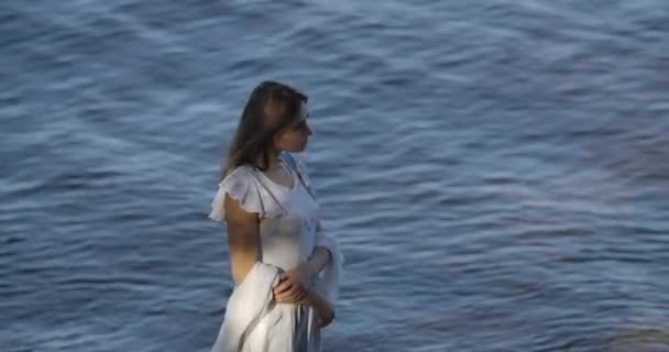 Vista superior de una hermosa mujer caucásica en vestido blanco claro de pie sobre el fondo de las olas del mar y el pensamiento. Chica atractiva pasar la noche de verano en la playa . — Vídeos de Stock