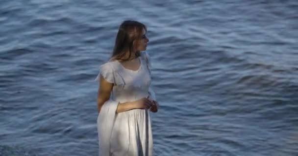 Droevige brunette Kaukasische vrouw in licht witte jurk staande op de achtergrond van de zee golven en denken. Aantrekkelijk meisje brengen zomeravond op het strand. — Stockvideo