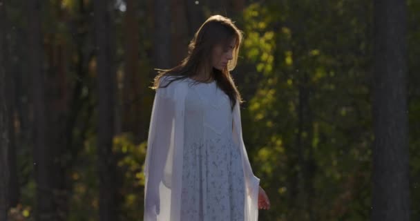 Urocza biała dziewczyna stojąca w słońcu w lesie i spoglądająca w bok. Piękna kobieta w jasnobiałej sukience spędza letni wieczór samotnie na świeżym powietrzu. — Wideo stockowe