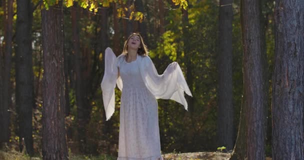 Vacker vit flicka som står i solljus i skogen, ringer någon och ler. Vacker kvinna i ljus vit klänning ha kul ensam utomhus. — Stockvideo