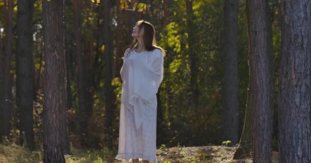 Charmante femme caucasienne enveloppée dans un châle blanc clair debout dans la lumière du soleil dans la forêt d'été et regardant autour. Belle fille appréciant calme soirée d'été en plein air . — Video