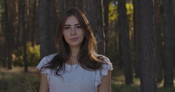 Portrait of a cute Caucasian brunette girl with brown eyes standing between trees in the summer forest. Charming woman in white dress posing to the camera and smiling. — ストック動画