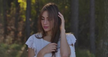 Yazlık ormanda ağaçların arasında duran kahverengi gözlü, düşünceli, Kafkas esmer bir kızın portresi. Beyaz elbiseli çekici kadın kameraya poz veriyor ve gülümsüyor..