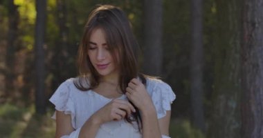 Yaz ormanında ağaçların arasında duran kahverengi gözlü, gülümseyen beyaz esmer bir kızın portresi. Beyaz elbiseli çekici kadın kameraya poz veriyor ve gülümsüyor..