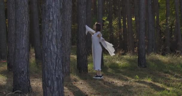 Menina caucasiana feliz correndo entre as árvores na floresta de verão e sorrindo. Mulher independente misteriosa em vestido branco claro se divertindo ao ar livre . — Vídeo de Stock