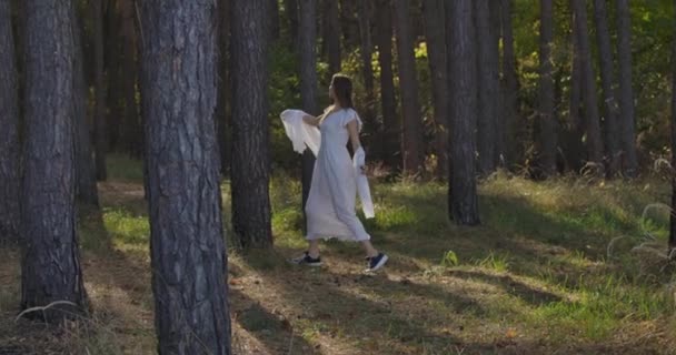 Happy Caucasian girl walking between the trees in the summer forest and smiling. Mysterious independent woman in light white dress having fun outdoors. — Stock Video