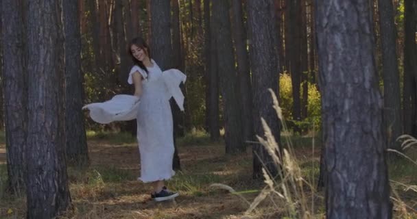Happy smiling Caucasian girl spinning around between the trees in the summer forest and smiling. Mysterious independent woman in light white dress having fun outdoors. — ストック動画