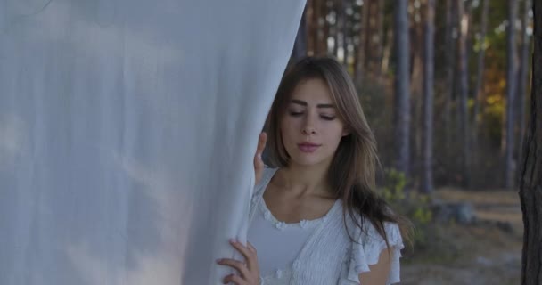 Portrait of a young beautiful Caucasian woman standing in the forest and looking around. Pretty girl holding white clothing hanging on the tree. — ストック動画