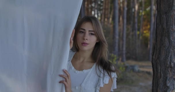 Porträtt av en ung vacker vit kvinna med bruna ögon som står i skogen och ser sig omkring. Söt flicka med vita kläder hängande på trädet. — Stockvideo