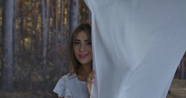 Ung positiv kaukasisk kvinna som kikar ut ur vita kläder och ler. Vacker flicka njuter av lugn sommardag i skogen. — Stockvideo